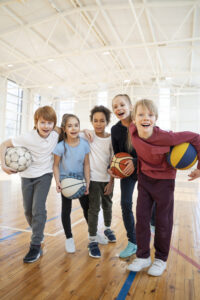 the group of five kids in the gym holding a basketball, soccer ball, and volleyball